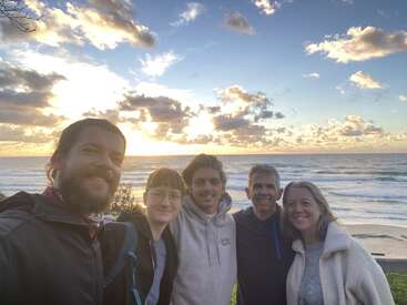 Cinq personnes se tiennent côte à côte, souriant à l'appareil photo, avec l'océan et un ciel de coucher de soleil spectaculaire, parsemé de nuages, en arrière-plan, créant une atmosphère chaleureuse et joyeuse.