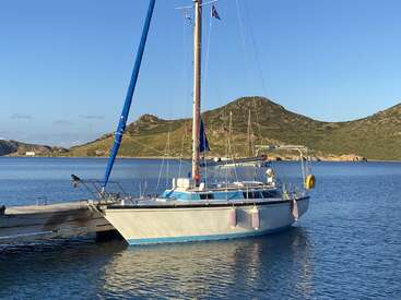 Das Bild zeigt ein weißes Segelboot mit blauen Verzierungen und einem hohen Mast, das an einem Steg in einem ruhigen Gewässer angedockt ist, umgeben von Hügeln und einem klaren blauen Himmel.