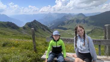 Two children sit on a wooden bench atop a scenic mountain, surrounded by green hills, wildflowers, and dramatic peaks under a bright, partly cloudy sky.