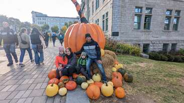 A family of four sits in front of a large decorative pumpkin, surrounded by various pumpkins and gourds, with people walking by in the background.