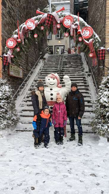 A family stands in snowy winter clothes before Bonhomme, under a festive archway with red decorations, smiling amid falling snow on decorated stone steps.