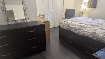 This image shows a tidy bedroom with a dark dresser, a mirror, a wooden desk and chair, a bed with drawers, and a standing lamp.