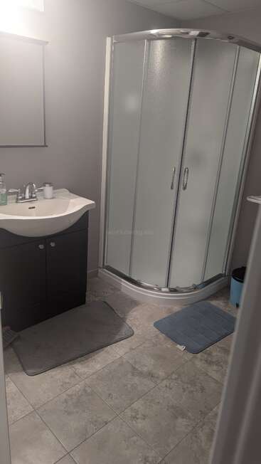 This image shows a clean bathroom with a corner shower, frosted glass doors, a sink with a dark cabinet, grey tiles, and two bath mats.
