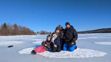 A imagem mostra uma família de quatro pessoas posando em um lago congelado, cercada por terreno coberto de neve e árvores, com um céu azul claro ao fundo.