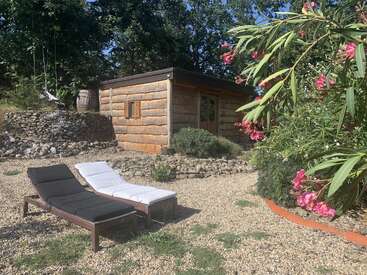 Uma cabana de madeira aconchegante fica entre árvores e pedras. Duas espreguiçadeiras estão sobre cascalho. Flores cor-de-rosa e vegetação exuberante dão cor ao cenário tranquilo do jardim.