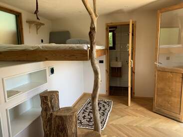 Esse quarto tem uma cama alta, detalhes em madeira rústica, degraus feitos de troncos de árvores, luz natural, um banheiro pequeno, prateleiras embutidas e uma decoração minimalista com tons terrosos.