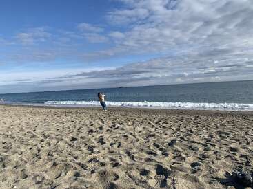 Uma pessoa corre alegremente por uma praia arenosa sob um céu parcialmente nublado. Ondas suaves quebram, criando uma cena costeira tranquila e relaxante.