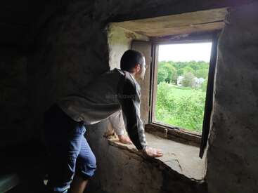 Um homem se inclina para fora de uma velha janela de pedra, contemplando a paisagem verdejante lá fora. A luz invade o ambiente, realçando o interior rústico. Um momento de paz.