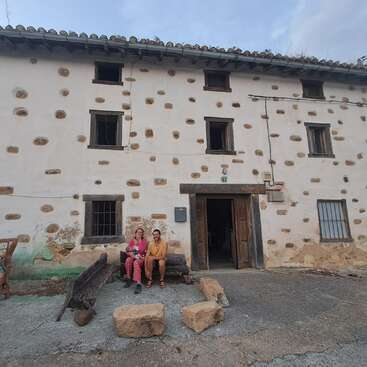 Um casal está sentado num banco em frente a um prédio rústico, caiado de branco, com detalhes em pedra, janelas de madeira e a porta aberta, sob um céu nublado.