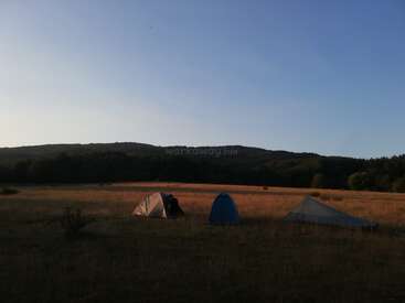 Três barracas estão montadas num campo aberto e gramado, sob um céu limpo ao pôr do sol, cercadas por colinas distantes e uma floresta densa, criando uma atmosfera tranquila.