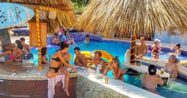 La imagen representa un entorno tropical con piscina y palmeras, en el que aparece un grupo de personas disfrutando en un ambiente relajado y tranquilo, rodeadas de exuberante vegetación.