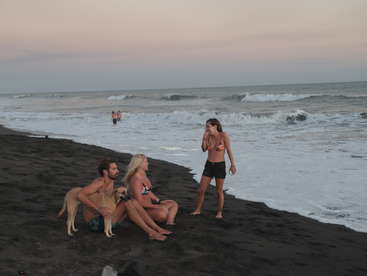 La imagen representa una serena escena playera con tres individuos, un perro y una perra, situados en la orilla, con las olas rompiendo suavemente en el borde del agua al fondo.