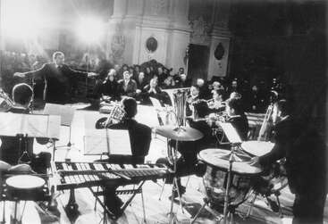Un director dirige una orquesta sentada que actúa en el escenario. Los músicos tocan varios instrumentos mientras el público observa atentamente en una sala de conciertos elegante y bien iluminada.