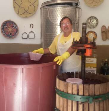 Un hombre con guantes amarillos y delantal trabaja con grandes recipientes, posiblemente haciendo vino, en una sala rústica decorada con cestas, platos y otros objetos tradicionales.