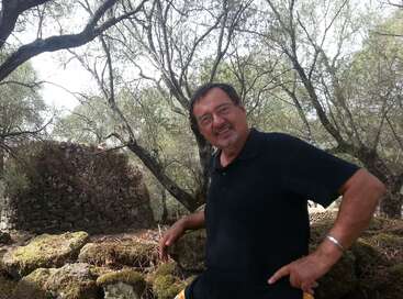 Un hombre con gafas y camisa negra sonríe mientras posa al aire libre entre rocas cubiertas de musgo, antiguas ruinas de piedra y árboles con extensas ramas al fondo.