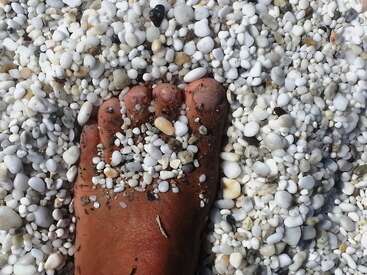 Un bronceado pie descalzo parcialmente cubierto de pequeños guijarros y piedras blancas, rodeado por una playa de guijarros lisos y redondeados de diversos tamaños y formas.