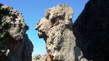 A large rock formation resembling a lion’s head stands between two cliffs under a bright blue sky, surrounded by rugged scenery and a few green plants.