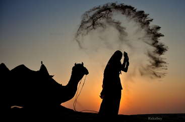 The image depicts a serene desert scene, featuring a camel and its rider, with the camel\'s humps and rider\'s clothing prominently displayed, set against a vast, arid landscape.