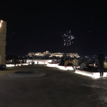 La imagen muestra un patio en una azotea por la noche, con el horizonte de la ciudad de fondo y fuegos artificiales explotando en el cielo. El patio está iluminado con luces.
