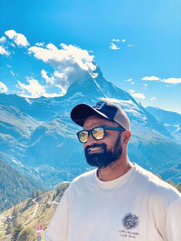A man wearing sunglasses and a cap smiles in front of a stunning mountain landscape, with clear blue skies and the iconic Matterhorn in the background.