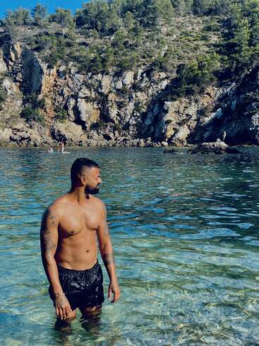 A shirtless man stands in clear, shallow water by a rocky coastline with lush greenery. Paddleboarders are visible in the background under a bright blue sky.