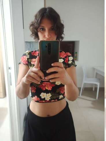 Uma mulher com cabelo curto e ondulado tira uma selfie no espelho. Ela está vestindo um top floral curto e uma saia preta, em pé em uma sala bem iluminada com decoração moderna.
