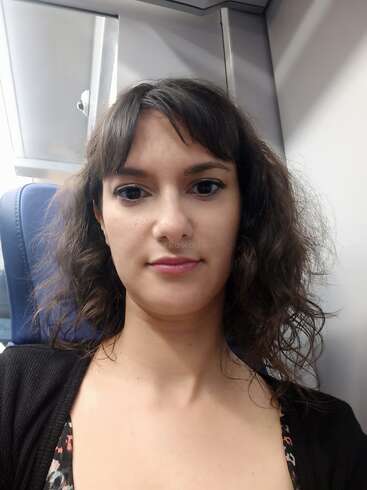 Uma mulher com cabelo castanho ondulado e maquiagem natural está sentada em um assento azul em um transporte público, provavelmente um trem, olhando diretamente para a câmera com uma expressão neutra.