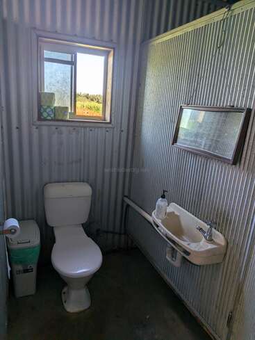 Il s'agit d'une petite salle de bains rustique avec des murs en tôle ondulée, des toilettes, une poubelle, un lavabo, du savon pour les mains, un miroir suspendu et une fenêtre sur laquelle sont posées des boîtes de mouchoirs.