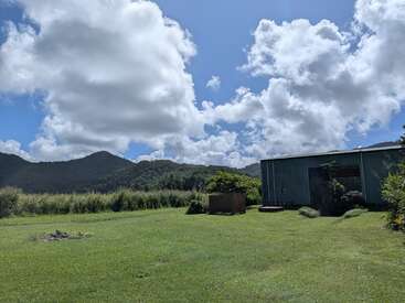 Un ciel bleu éclatant parsemé de nuages cotonneux au-dessus d'un champ verdoyant, des collines en arrière-plan et un hangar métallique sur la droite : une scène champêtre paisible.