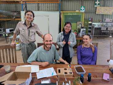 Quatre personnes, souriantes, sont réunies autour d'une table en bois, entourées de cahiers, d'échantillons de terre et de matériel scientifique, dans un atelier où l'on aperçoit des panneaux de premiers secours et du matériel de cuisine à proximité.