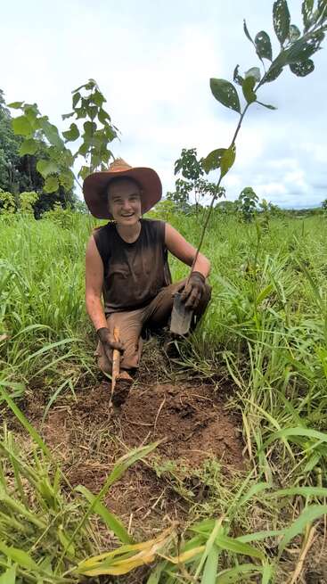 Une personne portant un chapeau à large bord et des gants est agenouillée dans les hautes herbes ; elle sourit en plantant un jeune arbre dans la terre fraîchement retournée, par une journée nuageuse.