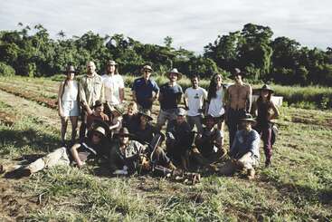 Das Bild zeigt eine Gruppe von 16 Personen, die für ein Foto auf einem von Bäumen umgebenen Feld posieren, wobei einige Hüte tragen und andere Werkzeuge in der Hand halten.