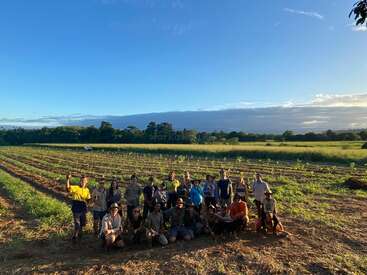 Un groupe de personnes pose ensemble dans un vaste champ agricole au coucher du soleil, célébrant le travail d'équipe et l'agriculture. Derrière elles s'étendent des rangées de cultures et un paysage verdoyant.