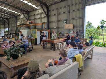 Un groupe de personnes est assis sur des canapés et des chaises dans un grand hangar ouvert et écoute attentivement une présentation. L'ambiance semble décontractée et conviviale.