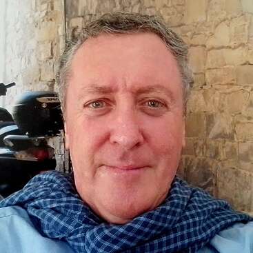 Ein Mann mittleren Alters mit hellen Augen und grauen Haaren lächelt in die Kamera; er trägt einen blau karierten Schal. Hinter ihm sind eine Steinmauer und ein Motorrad zu sehen.