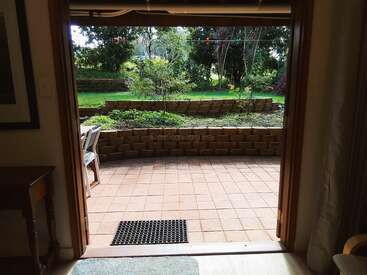 Vista desde el interior de una habitación hacia un patio de baldosas con un felpudo, dos sillas, un muro de contención, un jardín verde y árboles frondosos al fondo.
