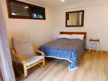 Esta imagen muestra un acogedor dormitorio con una cama de madera, ropa de cama a cuadros azules, un sillón claro, una mesita auxiliar, suelos de madera y un espejo de pared.