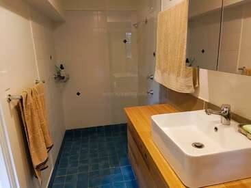 Este cuarto de baño cuenta con baldosas azules en el suelo, azulejos blancos en las paredes, un lavabo cuadrado de estilo moderno, un espejo grande, una encimera de madera, una ducha a ras de suelo y toallas de color beige colgadas con orden.
