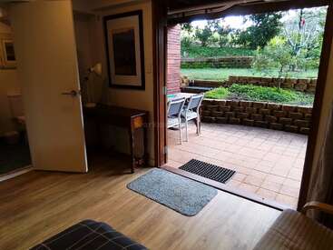 Una acogedora habitación que da a un patio con baldosas y a un jardín. En el exterior, una mesa con dos sillas; en el interior, suelos de madera, un escritorio, una lámpara y un cuadro enmarcado. Ambiente tranquilo.