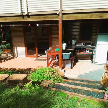 This image shows an outdoor patio area with a wooden table, chairs, a kitchenette, refrigerator, and greenery. Steps lead up from a small, lush garden.