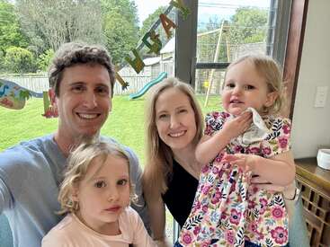 Eine vierköpfige Familie mit zwei kleinen Töchtern lächelt gemeinsam in einem Haus. Hinter ihnen steht bunte Geburtstagsdekoration und durch große Fenster ist ein grüner Hof zu sehen.