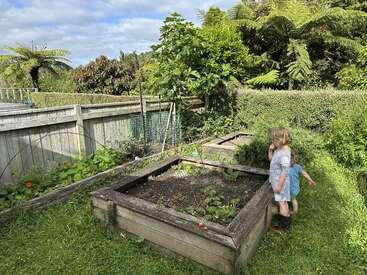Zwei kleine Kinder stehen neben Hochbeeten in einem üppigen, grünen Garten. Hohe Farne, Bäume, Hecken und ein Holzzaun schaffen eine ruhige Atmosphäre.