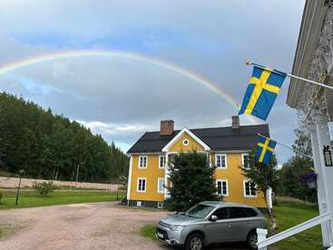 Un arc-en-ciel aux couleurs vives enjambe une maison jaune ornée de drapeaux suédois. Une voiture argentée est garée devant. Des arbres d'un vert luxuriant entourent ce cadre champêtre paisible.
