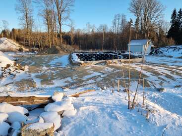 A snowy rural landscape with scattered logs, a small white shed, stacked black barrels, and bare trees, all bathed in bright winter sunlight. Quiet scene.