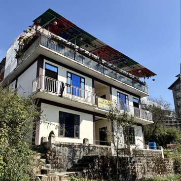 Un edificio de tres plantas con balcones y un colorido toldo en el tejado, rodeado de vegetación. Fondo de cielo azul, con escalones de piedra que conducen a la entrada, entorno tranquilo.