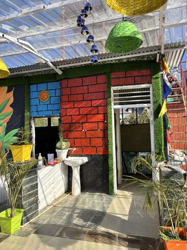 Colorida zona exterior con paredes con motivos de ladrillo, techo transparente, lámparas colgantes, macetas, un lavabo, campanas decorativas, luz solar y diversos adornos vibrantes que crean un espacio lleno de vida.