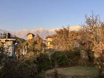La luz dorada del sol baña las casas rústicas y los árboles otoñales; las montañas nevadas se alzan majestuosas al fondo bajo un cielo azul despejado, creando una escena de pueblo serena y pintoresca.
