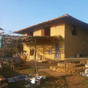 Das Bild zeigt ein rustikales, zweistöckiges Haus mit einer gelben Fassade und einem Ziegeldach, umgeben von einer Terrasse mit Gartenmöbeln und verschiedenen Gegenständen.