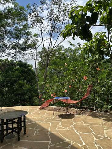 Un tranquilo patio al aire libre con suelo de piedra, una tumbona roja y mesas redondas, rodeado de frondosos árboles y flores rosas bajo un cielo parcialmente nublado.