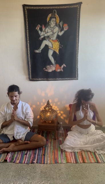 Dos personas están sentadas con las piernas cruzadas sobre una alfombra de colores, en postura de meditación, con las manos juntas en señal de oración, y entre ellas hay un tapiz de Shiva y una lámpara encendida.